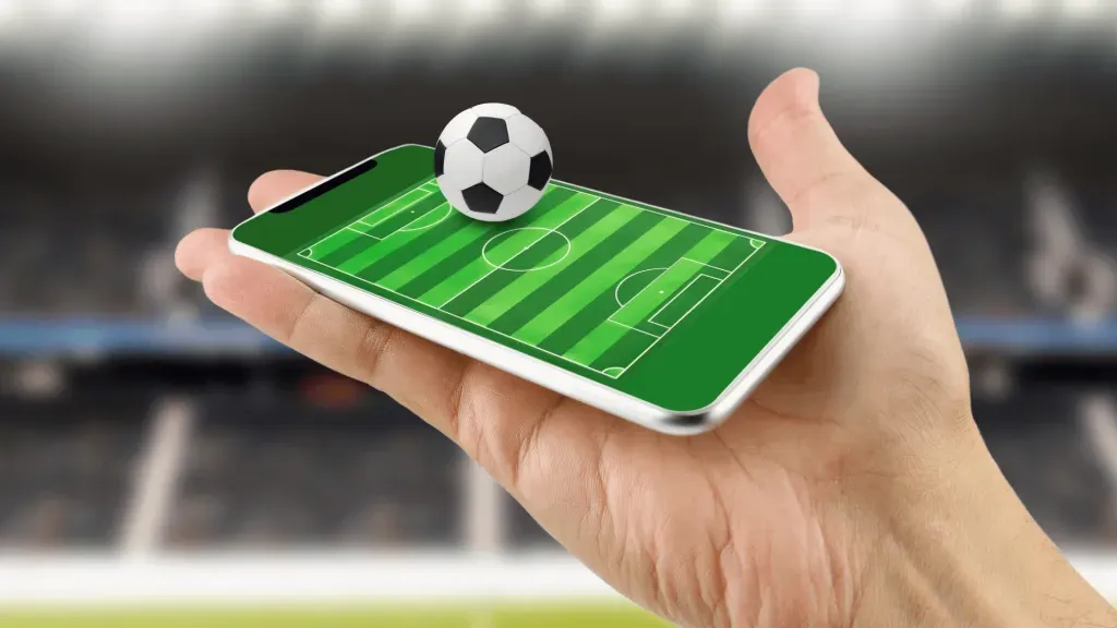 Imagem gráfica de mão segurando smartphone com projeção de campo de futebol na tela e a clássica bola branca a preta em 3d.