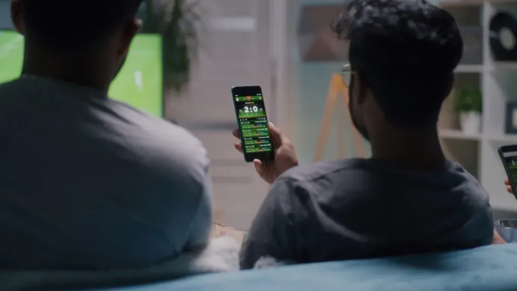 Dois homens lado a lado assistindo partida de futebol na TV. Um deles está segurando um smartphone aberto em um app de casa de apostas.