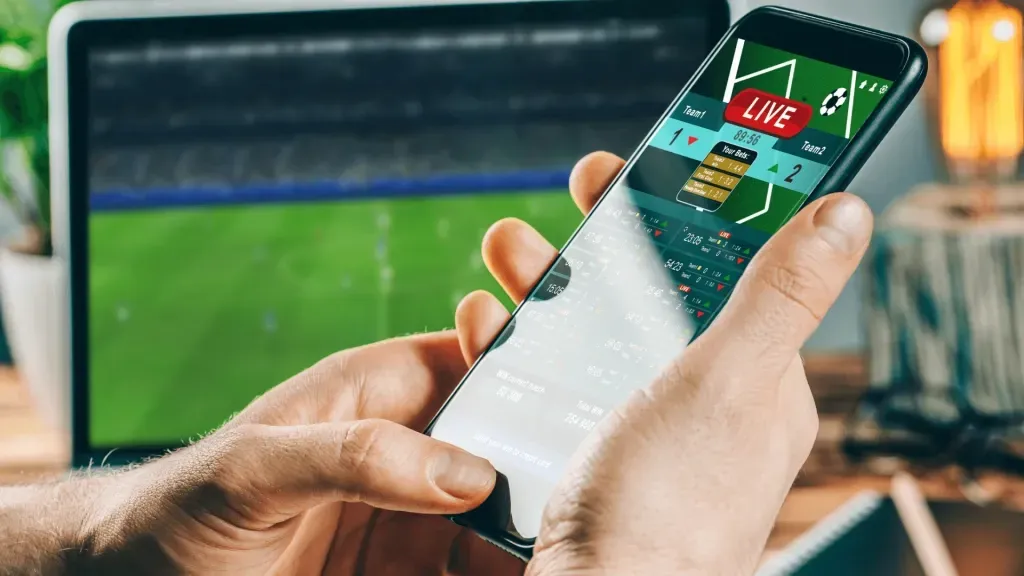 Mãos segurando smartphone aberto em casa de apostas, contra fundo de uma TV ligada em uma partida de futebol, representando os melhores apps de apostas do Brasil.